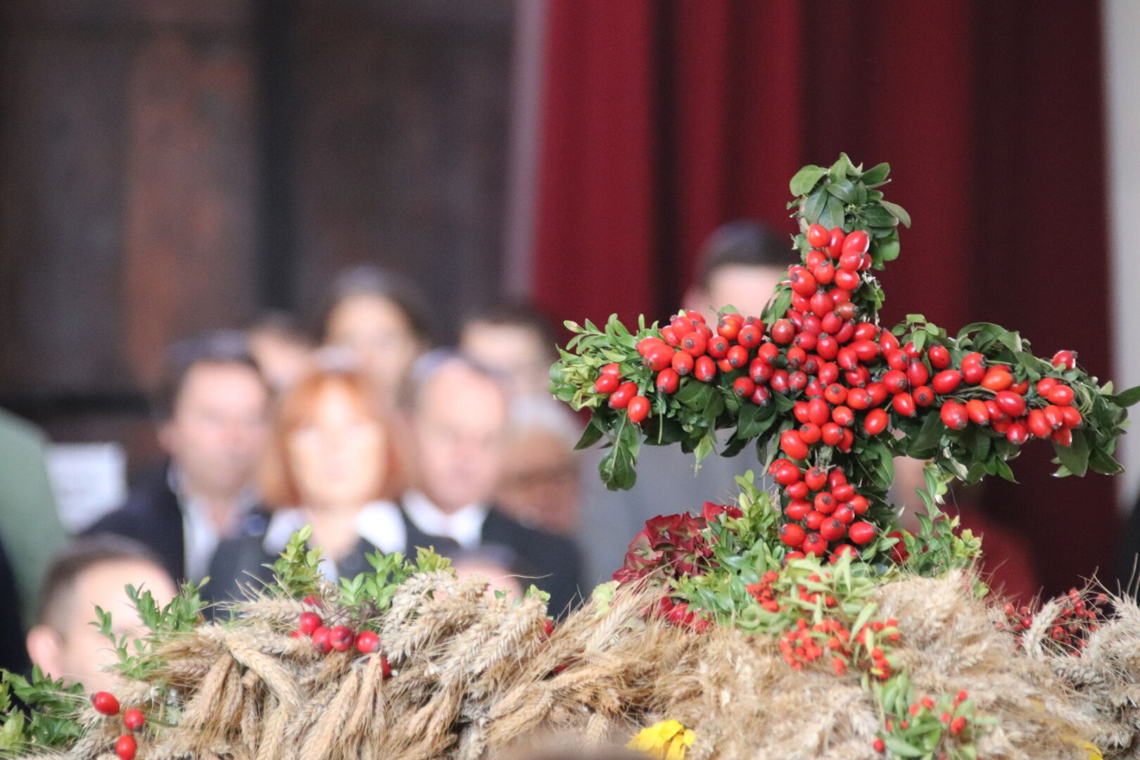 Ein Kreuz aus vielen roten Hagebutten und grünem Buchsbaum als Spitze auf einer Erntekrone, deren Getreide-Ähren am unteren Bildrand sichtbar sind. Im Hintergrund sind unscharf einige Menschen zu sehen, gekleidet hauptsächlich in grau und schwarz; weiße Krägen blitzen hervor.